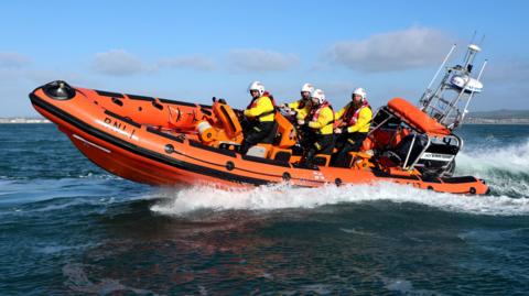Five RNLI rescuers in an orange RNLI boat at sea. They are wearing helmets. It is a sunny day.