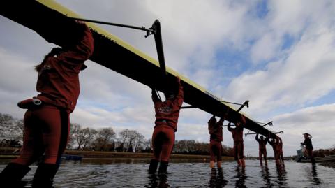 Seven women are holding a rowing boat above their heads. They're wading into a river as they carry the boat. They're wearing matching sports kits, which show they're rowers for Oxford Brookes rowing team. The sports jackets and trousers are red. The sky is blue and cloudy. 