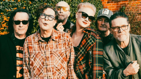 The band pose outside with a wall and greenery behind them. Three have sunglasses on and two are wearing checked shirts with another in a black jacket and a fourth member in a green jacket. Two others are obscured in the photo.