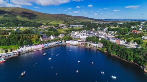 An aerial image of Portree showing boats in the bay and house, some colourfully painted pink and blue, and set among trees.