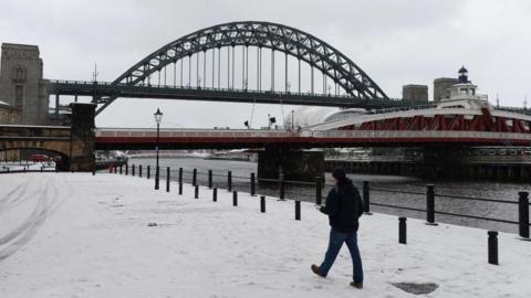 Man in black jumper and black beanie walking on snow covered quayside in front of green Tyne Bridge and red Swing Bridge in Newcastle