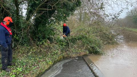 A worker wearing muddy black trousers, a blue jumper, black gloves and an orange helmet watches another similarly-dressed worker using a chainsaw to cut up a tree which is blocking a canal.