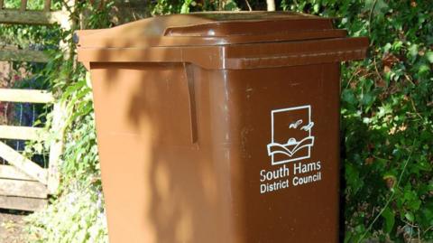 A brown wheelie bin with the words 'South Hams District Council' printed on the front in white writing, with an image of a sunset and birds.