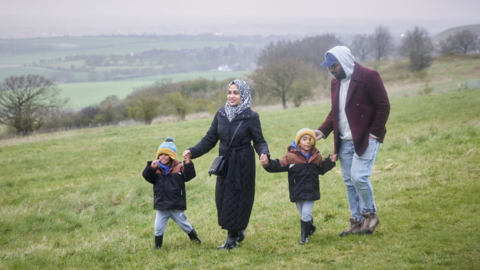 A woman, a man and two boys aged about four and five walking at Dunstable Downs. The boys are wearing matching dark coats, grey joggers, black wellies and knitted hats. The woman is holding each boy by the hand and is wearing a black full length coat and black and white head scarf. The man is wearing pale blue jeans, a brown jacket, a grey hooded top with the hood up over a blue baseball cap