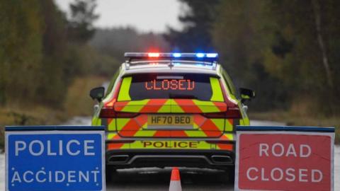 Accident sign and road closed sign either side of the back end of a police car with lights flashing on its roof.