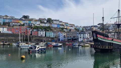 A number of small boats sit idly in Brixham harbour against a backdrop of small, colourful houses 