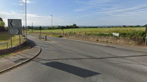 Leigh Crossroads as seen on a sunny day with road signs and a low stone border wall on a sunny day with blue skies overhead
