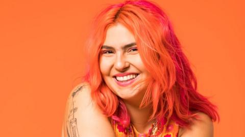 Ruby Rare, a young woman with dyed orange and pink hair, smiles at the camera. Ruby wears pink eyeshadow and lipstick and wears her hair loose around her shoulders. Her arms are folded across her front, revealing a fine-line tattoo on her right shoulder and down her arm. She wears a sleeveless orange and pink top and is photographed in front of an orange background,