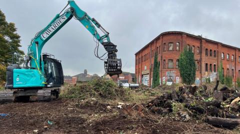 Derelict site Friar Gate Goods Yard in Derby appears in the background of the image with a blue digger in the forefront. The digger is working to remove overgrown areas around the site.