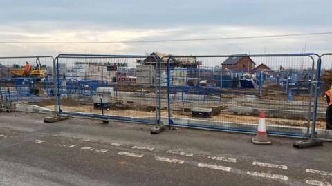Fencing surrounds a works site where a central reservation and pedestrian crossing is being built. In the background is machinery and houses under construction.