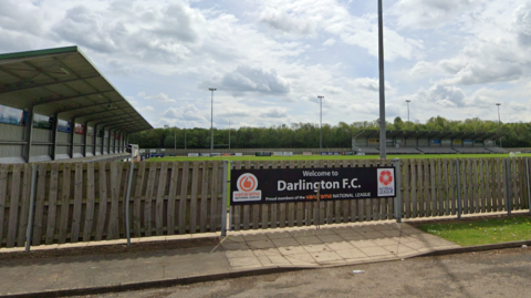Blackwell Meadows is a football pitch with a wooden fence surrounding it. A sign that reads 'Welcome to Darlington FC' with various sponsorships can be seen on the fence in front. Spectator stands are in the distance.