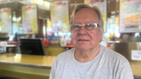 A man with white hair, wearing a grey t-shirt, sits at the bar of an ice cream parlour.