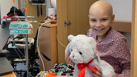 Poppi in a hospital bed cuddling a large teddy bear. She is wearing red pyjamas with a white pattern and is smiling. She has lost her hair because of her treatment. There is a medical machine and a chest of drawers next to the bed.