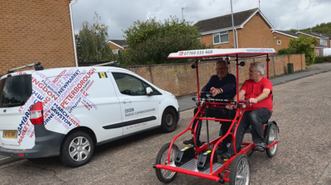 Two men ride on a red surrey bike, one in a blue fleece, the other in a red t-shirt, the machine has four-wheels and two sets, it has a white canopy on top it is being ridden on a street with a white BBC van parked on the side on the road.
There are houses visible in the background.
