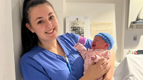 A woman with long dark hair in a high ponytail wearing blue scrubs holds a small crying baby in a pink onesie and a blue hat.