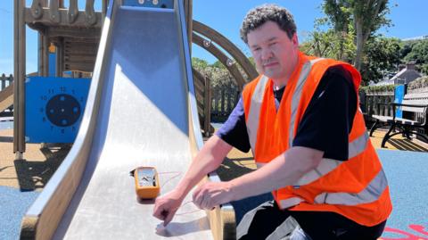 A man in an orange high vis vest holds a thermometer to the bottom of a silver, metal slide, while looking straight-faced at the camera. 