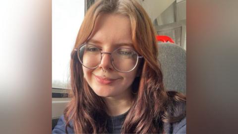 Corinna Baker. She has red hair, is smiling and is wearing a pair of large circular spectacles. She has a nose ring and stud through her lip