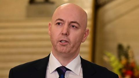 A bald man with short red stubble looks to the left of the frame. He wears a balck suit and purple and navy tie. He is standing in the Great Hall at Stormont in front of cream stairs, addressing the press 