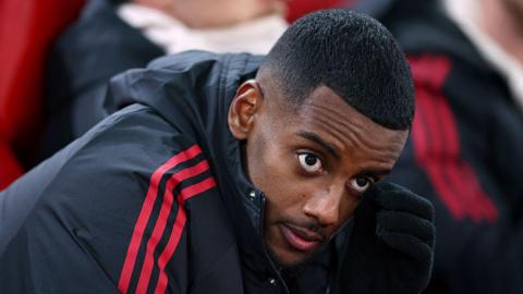 Alexander Isak looks on from the bench in a zip-up dark jacket with red stripes before the Champions League game between Liverpool and PSV Eindhoven at Anfield on 26 November, 2025