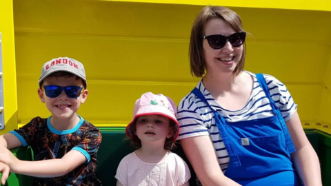 Nicola with Oscar and Ffion. The children are wearing sunhats and t-shirts and Nicola is wearing blue dungarees over a blue and white striped t-shirt. All are smiling.