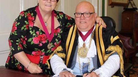 A man sitting down in mayoral garb and wearing glasses.