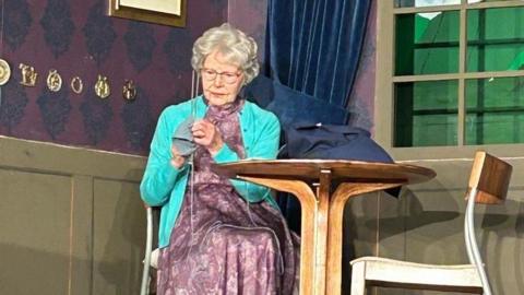 A woman sits on a chair at a small table on stage. She has grey hair and glasses, and is holding a needle and some material