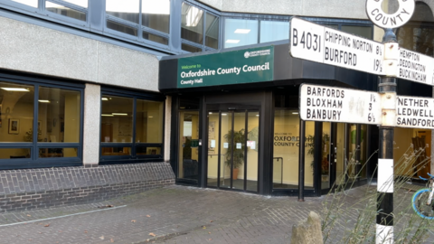 Oxfordshire County Council's County Hall in Oxford. The picture shows the front doors of the building, with a green sign above that says "Welcome to Oxfordshire County Council County Hall". There is a black and white sign to the right of the picture pointing out distances to other places in Oxfordshire.