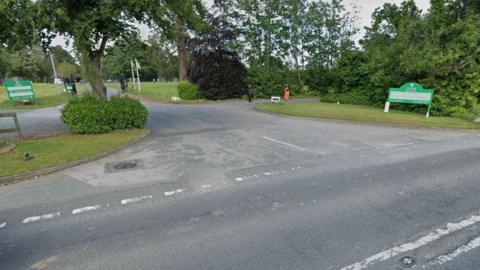 A street view image showing the entrance to Stover School from the A382. A green sign for the school with directions to the visitor car park is on a grass verge by the entrance. Two people, one of whom is wearing orange high-vis trousers, are having a conversation in the distance. A number of trees, shrubs and playing fields are dotted around the area.