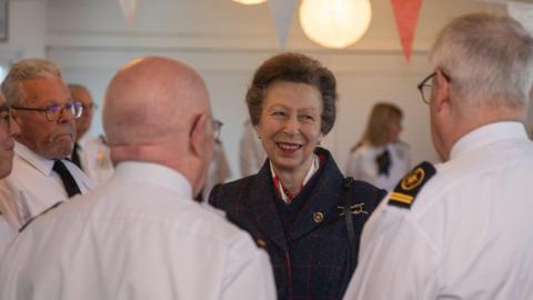 Princess Anne with brown hair is facing the camera and smiling while looking off to the right. She is wearing a dark blue coat. Three elderly men wearing white uniform shirts are facing hair