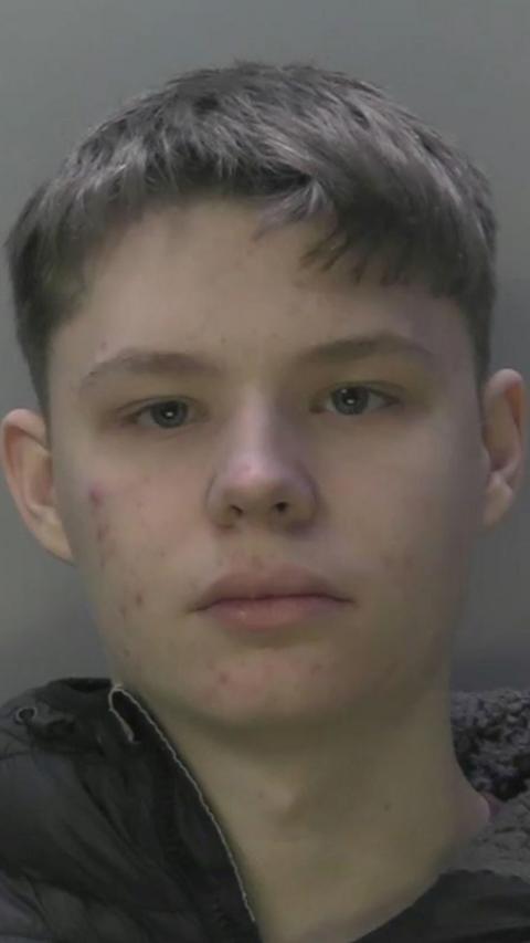 A mugshot of a boy with short brown hair and a black coat.