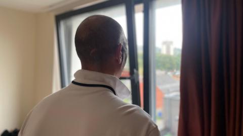 A man with a shaved head looks out of a window which is only narrowly open