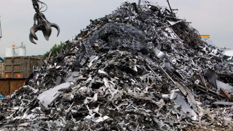 A huge pile of silver scrap metal, with a metal crane claw coming down towards it, to grab it.