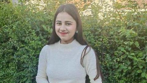 Harshita Brella smiles at the camera as she is photographed outside. There is a large green hedge behind her. She is wearing a beige polo-necked jumper and has long dark hair. She is wearing earrings. 