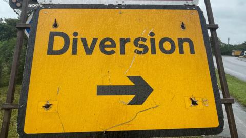 A close-up of a generic image of a yellow road sign saying diversion in black, with an arrow underneath pointing to the right. It is on a pavement beside a road. A lorry can be glimpsed turning to the right in the far distance. 