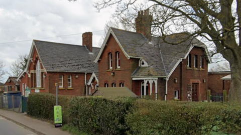 A Google Maps streetview image taken from the outside Bucklesham Primary School. It includes the main school building, in front of which there are green bushes and a tree. 