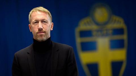 Graham Potter looks into the camera as he stands in front of a board displaying the badge of Sweden's football team.