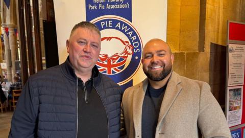 Alexander Melling stands to the right of another man under a advertising hoarding for the British Pie Awards. 