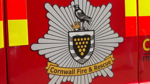 The image shows the emblem of Cornwall Fire & Rescue with a banner and red with yellow and red reflective stripes on either side.