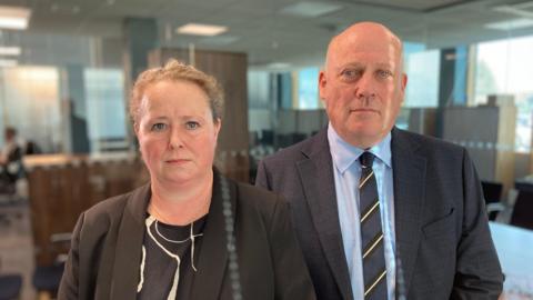 Two members of police staff look at the camera with a neutral expression. The woman on the left has fair hair and wears a black jacket over a black and white top. The man on the right has a shaved head and is wearing a dark suit and blue tie. 