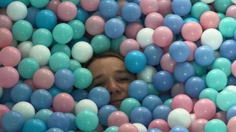 A woman's face seen popping amid dozens of pastel coloured balls in a ball put