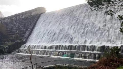 Water rushing down a dam. The structure is large and is made of stone. Water is fast flowing. Trees are next to the dam.
