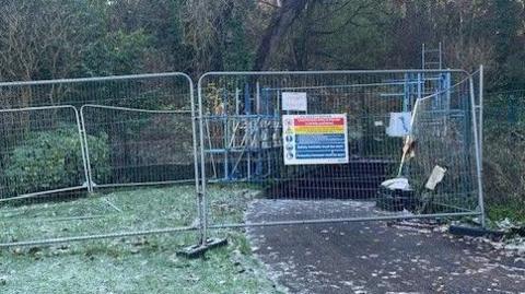 Grey fencing with a sign surrounds the Coleman's Field bridge. The grass and the leaves on the path are covered in frost. 