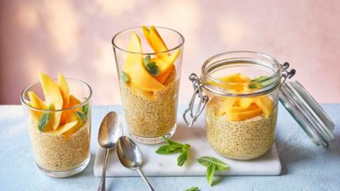 Three glasses of mango lassi chia pudding topped with slices of mango and mint on a white tray with two teaspoons placed next to them.