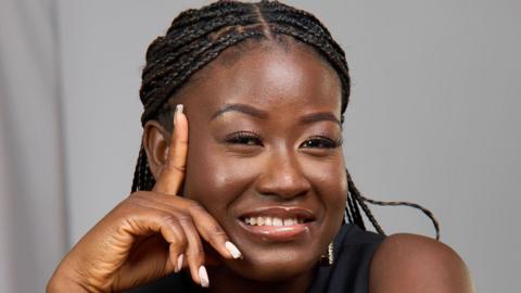 Woman smiles leaning her hand against her head. She has braids and wears a black top.