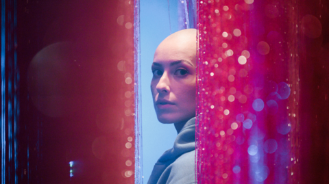 Emily Glaze is turning to look at the camera side-on from between pink glittery curtains. She has a bald head and is wearing a light blue hoodie.