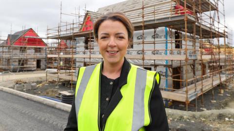Liz Kimmins stands in front of a house currently under sconstruction. It is covered by scaffolding. Kimmins is wearing a high viz yellow jacket and black blouse. She is smiling and has short brown hair