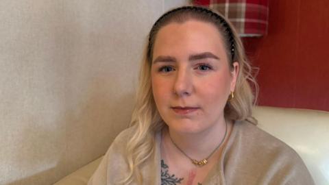 A woman looking at the camera with a neutral expression. She has blonde curly hair which is pushed away from her face with a black headband. She's wearing a beige jumper. An intricate tattoo across the top of her chest and a red vertical scar can be seen. 