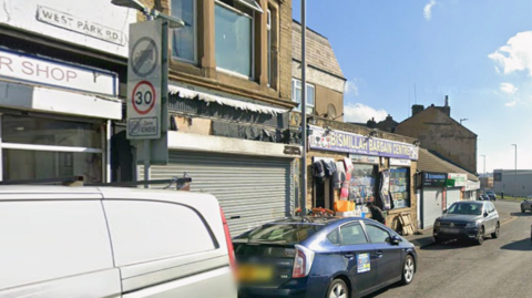 Cars and vans parked up at the side of a road in front of a row of shops.
