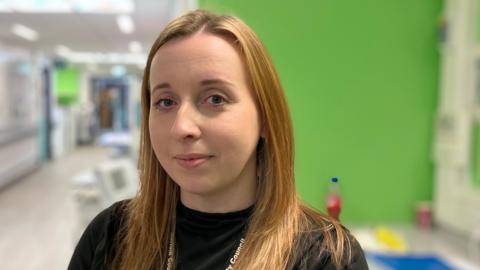 Abbey Cummings has long dark blonde hair and is wearing a black T-shirt and a lanyard. She is standing next to worktop, which is on the left, and has a water bottle and other items on it.