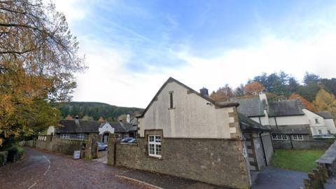 Forestry England buildings at Grizedale. It is a large construction of primarily two-stores and surrounding a courtyard. One of the buildings appears to be a converted stable block with central arch. A country lane snakes into the distance with mature trees at the side. In the background a large hill riases, covered in forest.